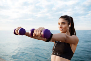 Woman performing outdoor dumbbell strength training by the water, representing active lifestyle habits that support metabolic health, weight management, and overall wellness.